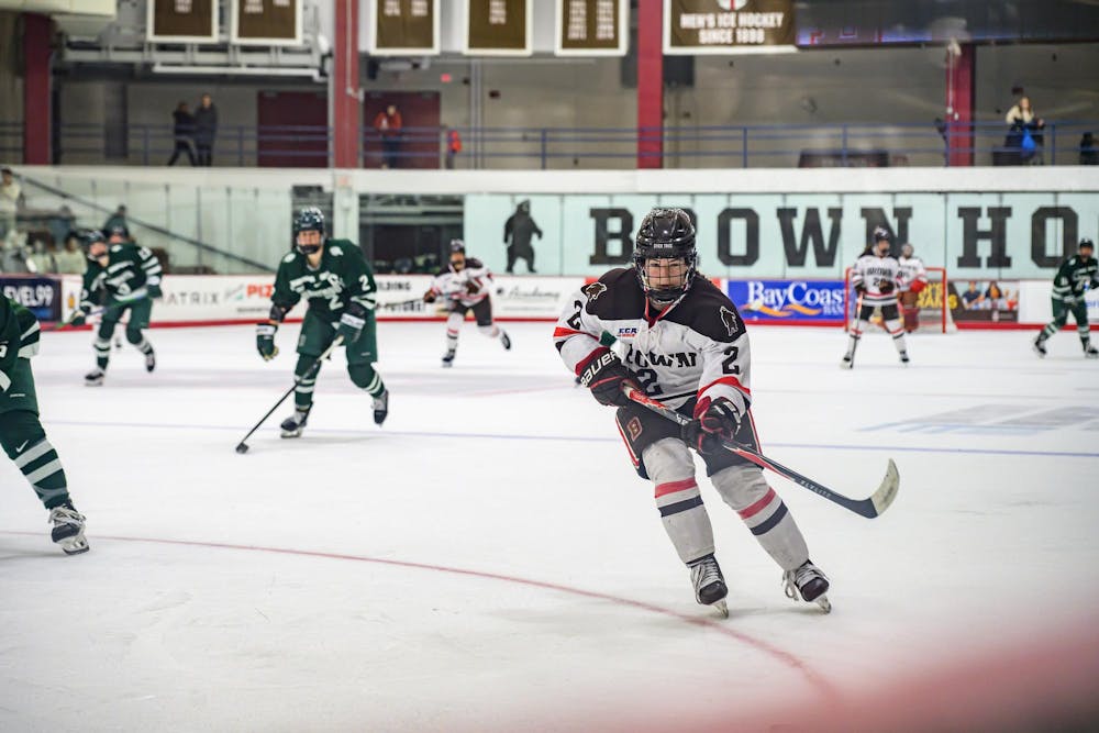A photo of a Brown University hockey player holding their hockey stick while skating on ice towards a sliding puck with other players in the background. 
