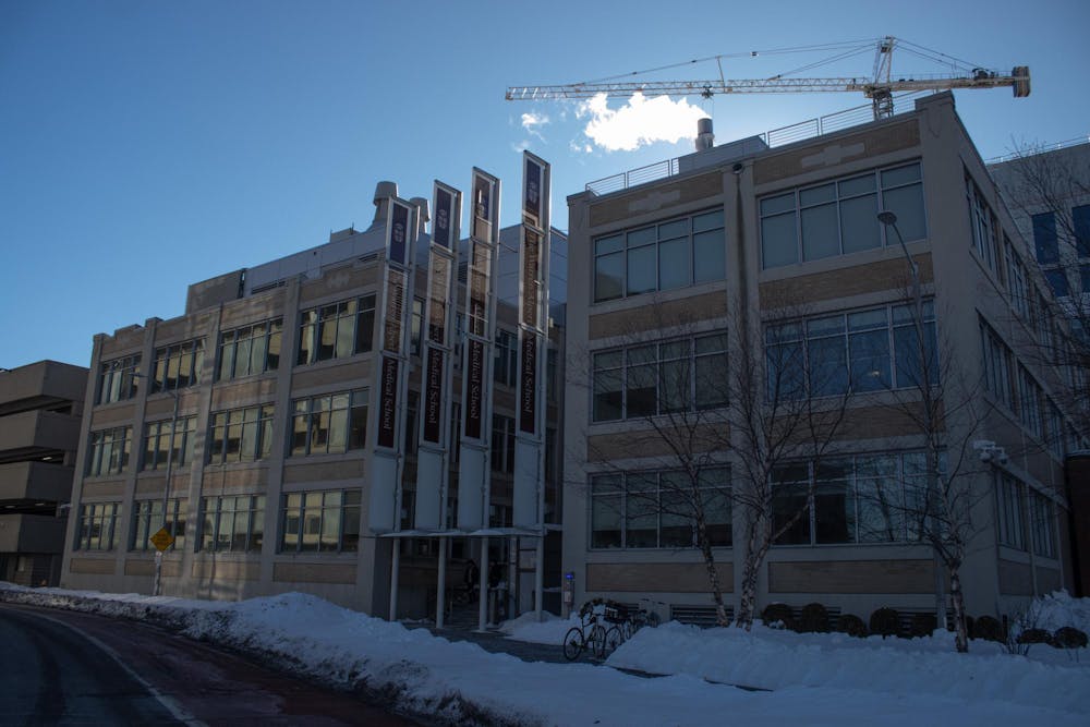 Photo of a long, rectangular building with large windows and four vertical brown banners displaying the words “Warren Alpert Medical School” in white text and the Brown University logo at the top. Some small piles of snow are in front of the building with a few bikes in front and on the bike rack.


