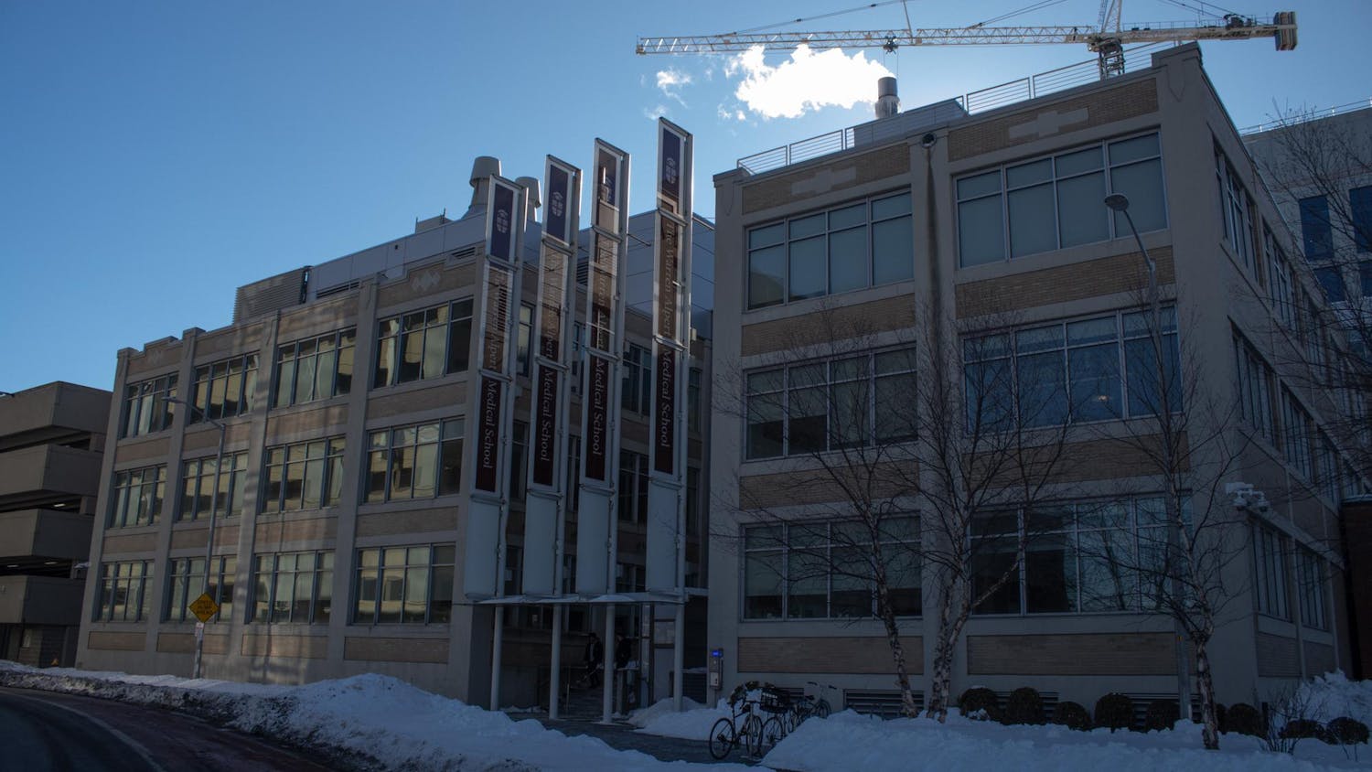 Photo of a long, rectangular building with large windows and four vertical brown banners displaying the words “Warren Alpert Medical School” in white text and the Brown University logo at the top. Some small piles of snow are in front of the building with a few bikes in front and on the bike rack.