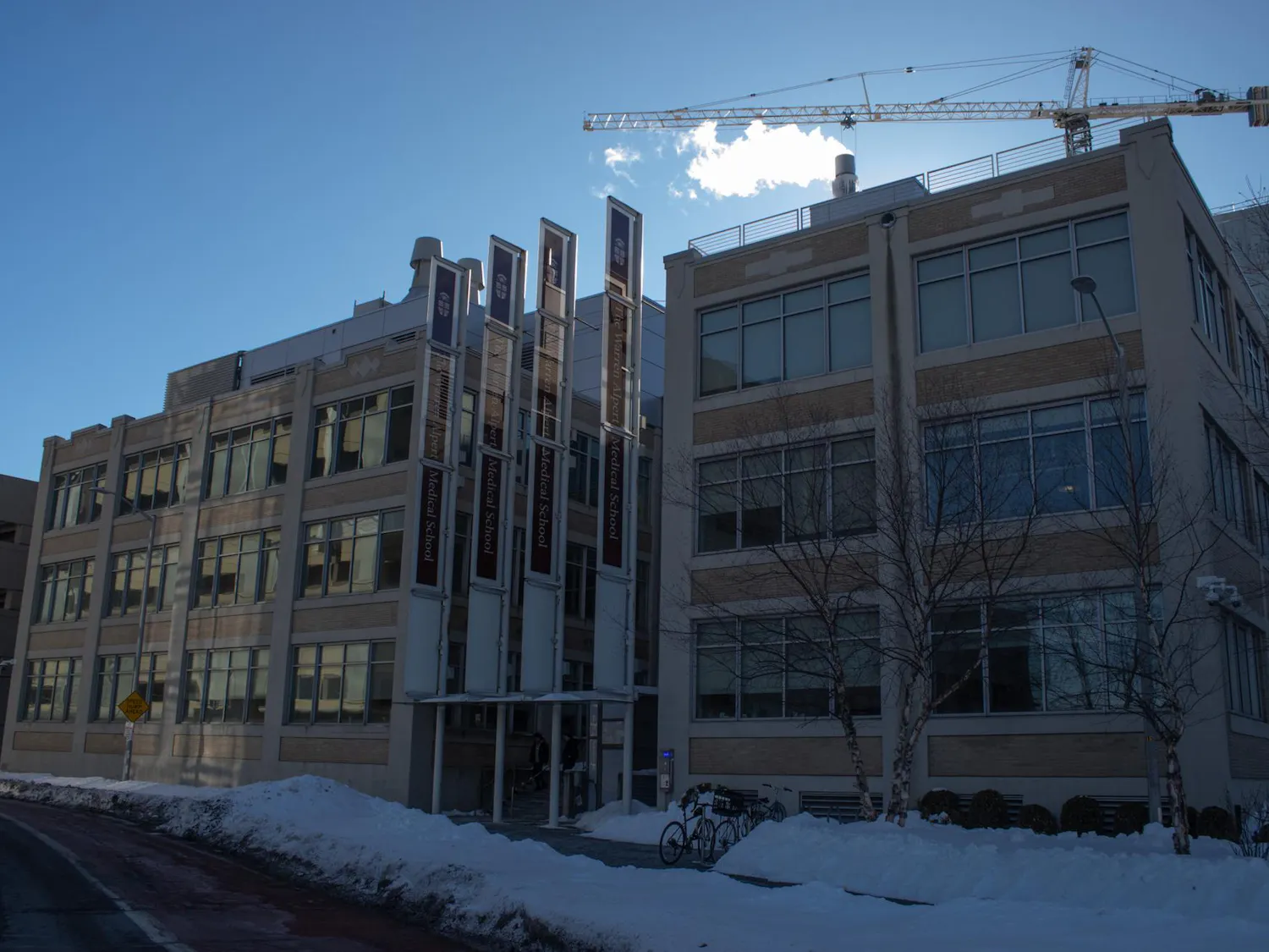 Photo of a long, rectangular building with large windows and four vertical brown banners displaying the words “Warren Alpert Medical School” in white text and the Brown University logo at the top. Some small piles of snow are in front of the building with a few bikes in front and on the bike rack.