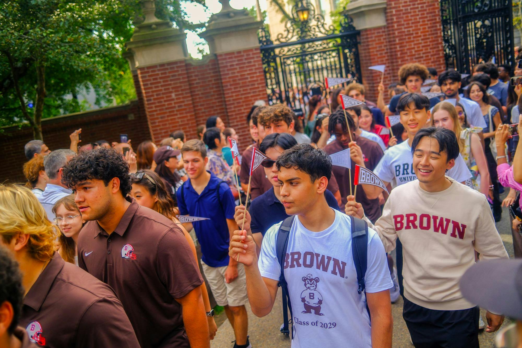 A photo showing a crowd of students walking left towards the camera holding small flags that read "Brown." Several students wear Brown University apparel. In the foreground, one student with dark hair looks to the left while another smiles behind him.