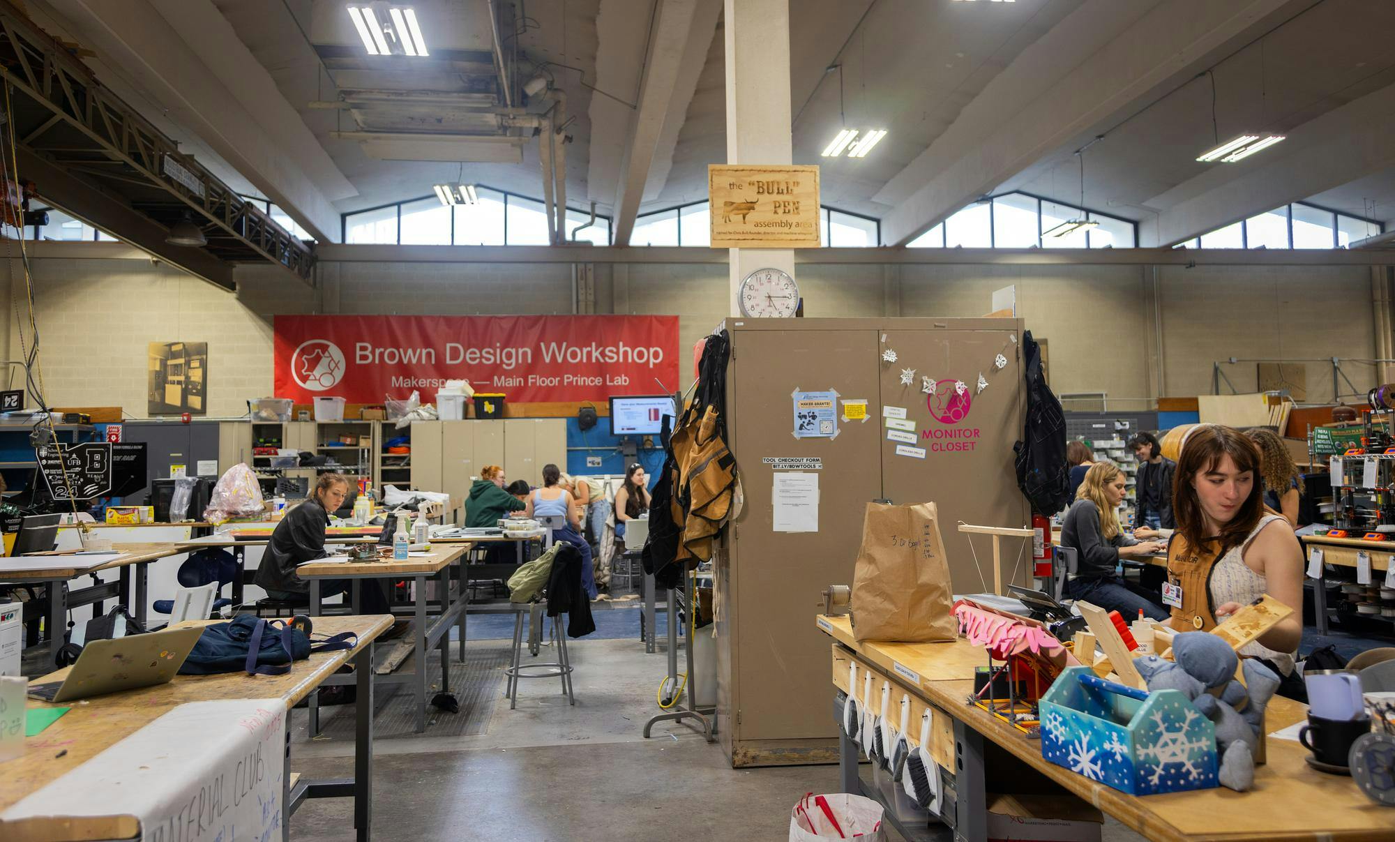 A picture of the Brown Design Workshop. Students can be seen sitting at numerous wooden workbenches scattered throughout the space.