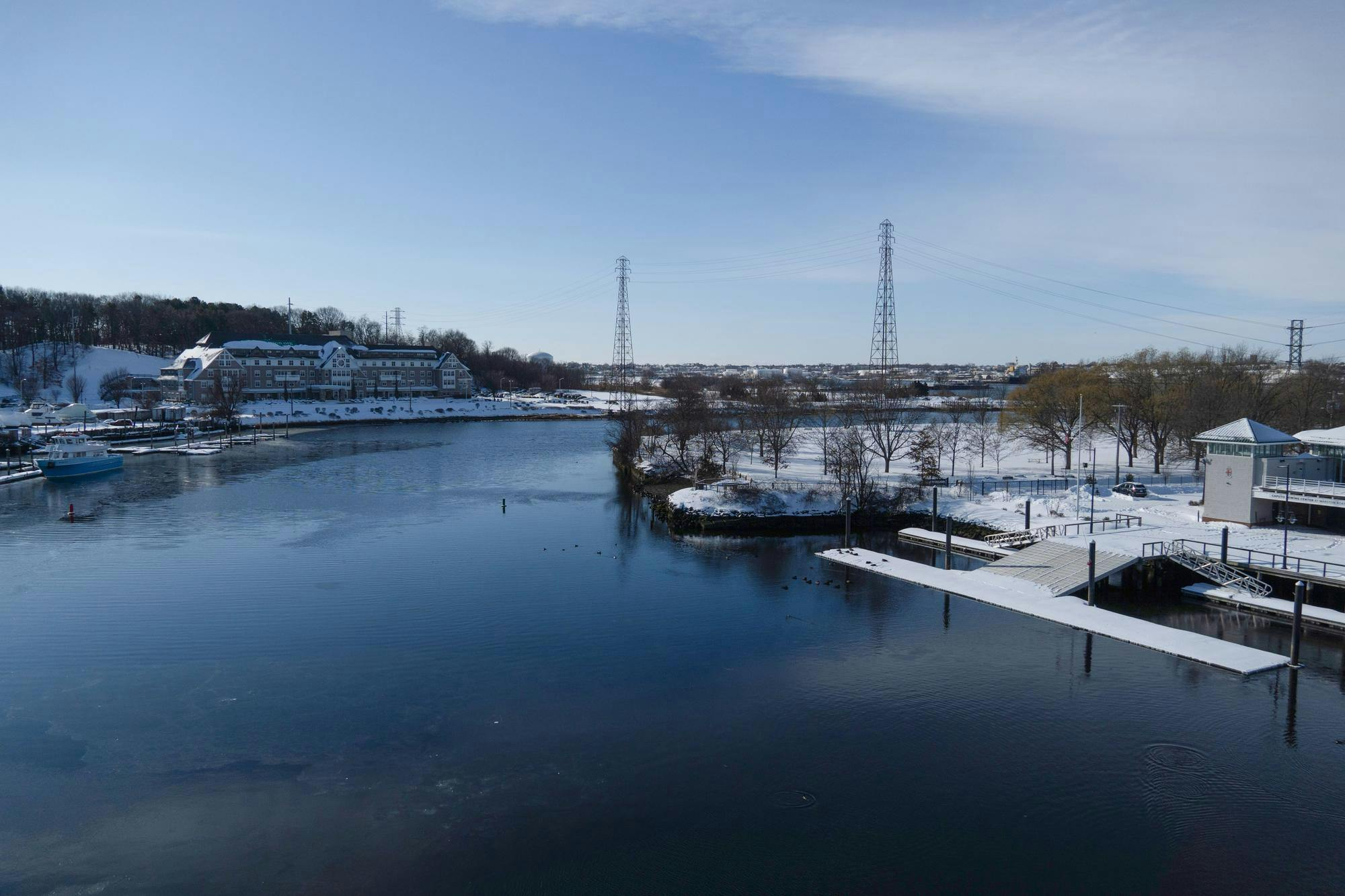 Landscape view of the Providence River in the winter with a snow-covered dock in the foreground, building in the middle ground and two power lines in the background. 
