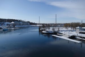 Landscape view of the Providence River in the winter with a snow-covered dock in the foreground, building in the middle ground and two power lines in the background. 