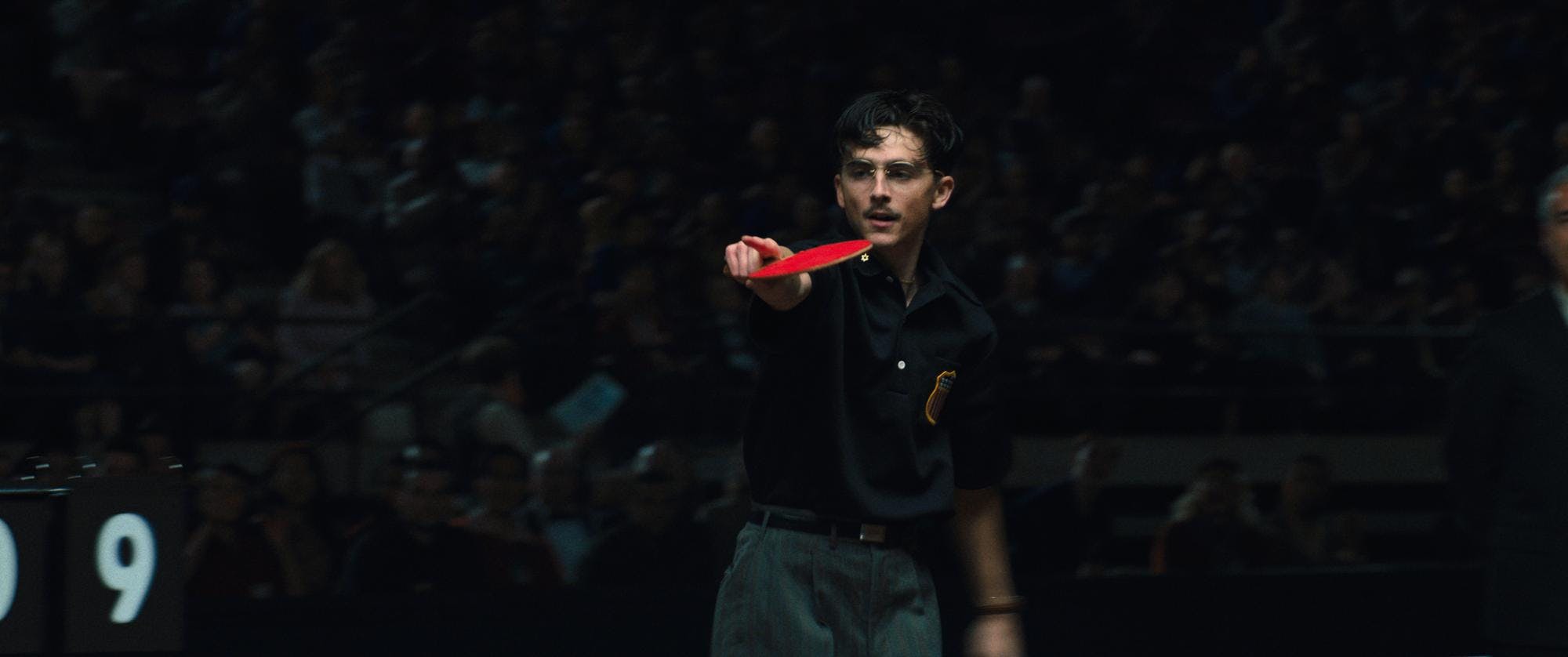 A man in a black shirt, grey pants and glasses holds a red ping pong paddle while pointing. 