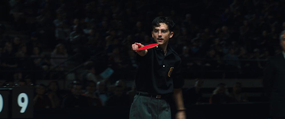 A man in a black shirt, grey pants and glasses holds a red ping pong paddle while pointing. 