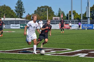Photo showing a Brown soccer player wearing a white uniform dribbling a soccer ball as a Cornell soccer player wearing a black uniform runs up behind him. 