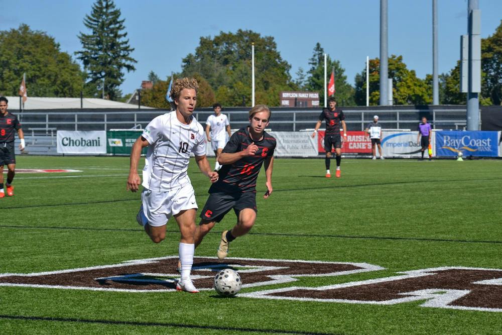 Photo showing a Brown soccer player wearing a white uniform dribbling a soccer ball as a Cornell soccer player wearing a black uniform runs up behind him. 