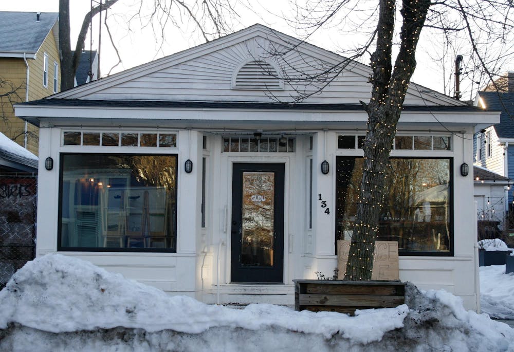 A photo of the front of a white house with large windows showing stacked chairs inside, “Glou” labeled on the door, and a “Garage Sale” sign leaning against the outside wall. 