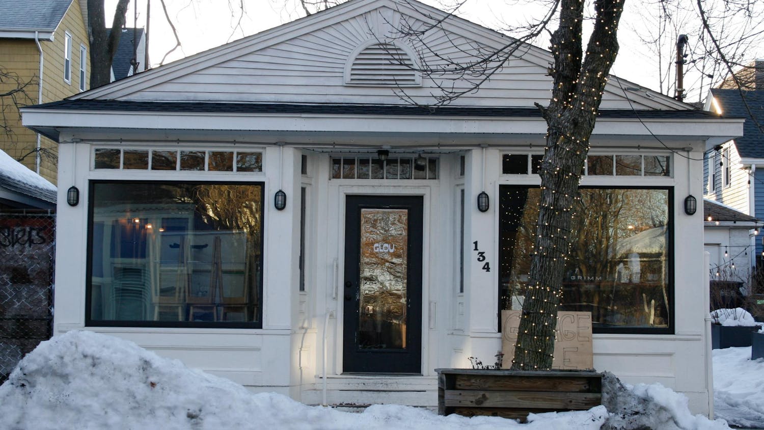 A photo of the front of a white house with large windows showing stacked chairs inside, “Glou” labeled on the door, and a “Garage Sale” sign leaning against the outside wall.