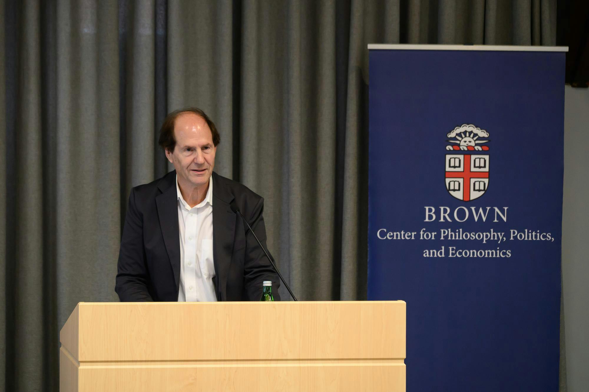 Photo of a man wearing a black suit jacket and white collared shirt standing at a podium. There is a blue sign behind him with the Brown insignia and the words “Center for Philosophy, Politics, and Economics”.