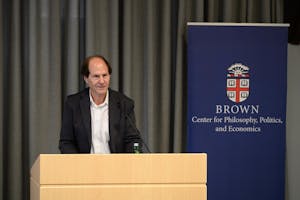Photo of a man wearing a black suit jacket and white collared shirt standing at a podium. There is a blue sign behind him with the Brown insignia and the words “Center for Philosophy, Politics, and Economics”.