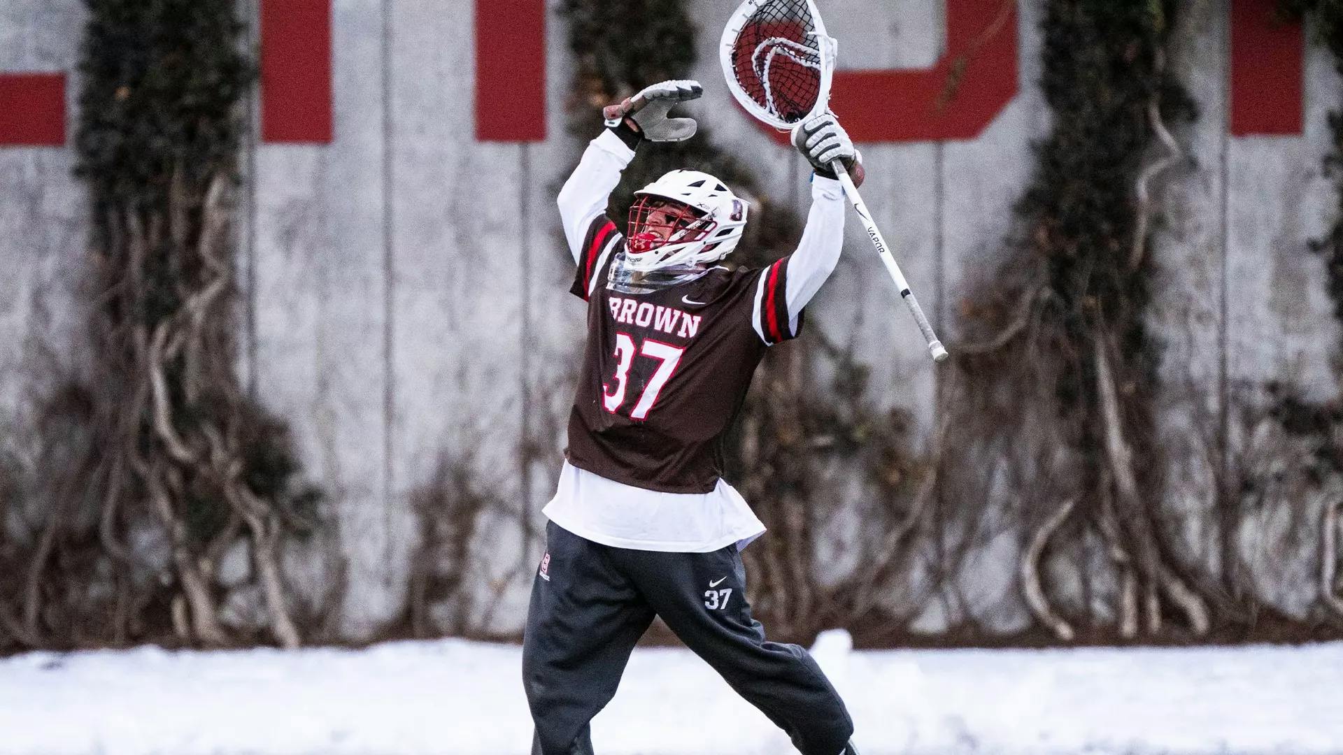 Photo of Brown Men’s Lacrosse player jumping, lifting his arms and lacrosse stick in the air.