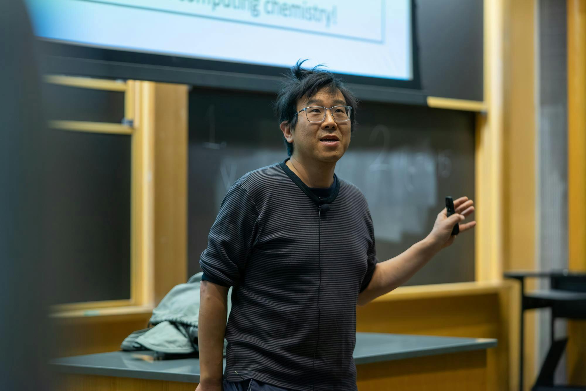A man with glasses and a striped long-sleeve t-shirt lectures in the front of an auditorium.

