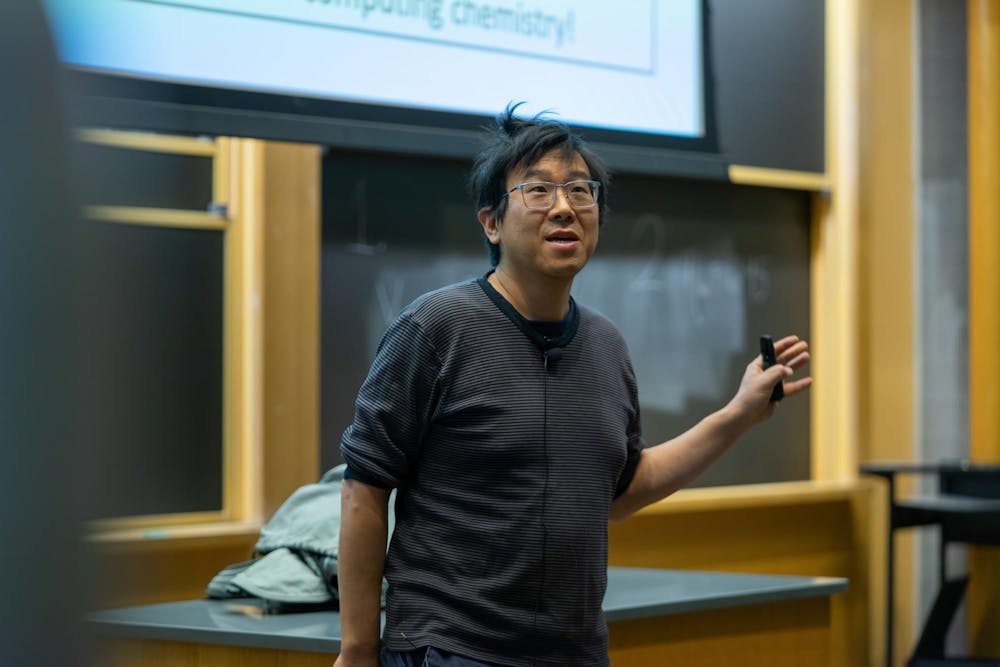 A man with glasses and a striped long-sleeve t-shirt lectures in the front of an auditorium.

