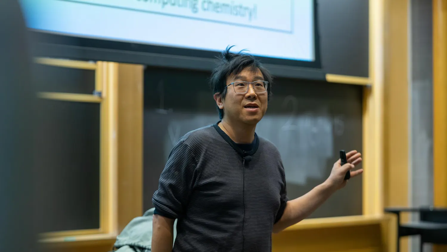 A man with glasses and a striped long-sleeve t-shirt lectures in the front of an auditorium.
