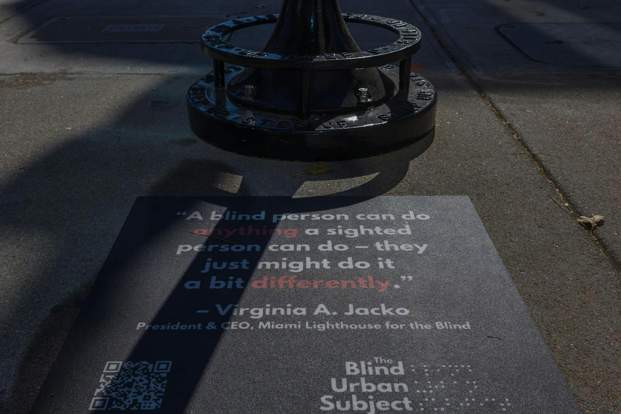 A sign on a sidewalk that reads, "'A blind person can do anything a sighted person can do — they just might do it a bit differently.' — Virginia A. Jacko, President & CEO, Miami Lighthouse for the Blind." The Blind Urban Subject logo is featured at the bottom of the sign. 