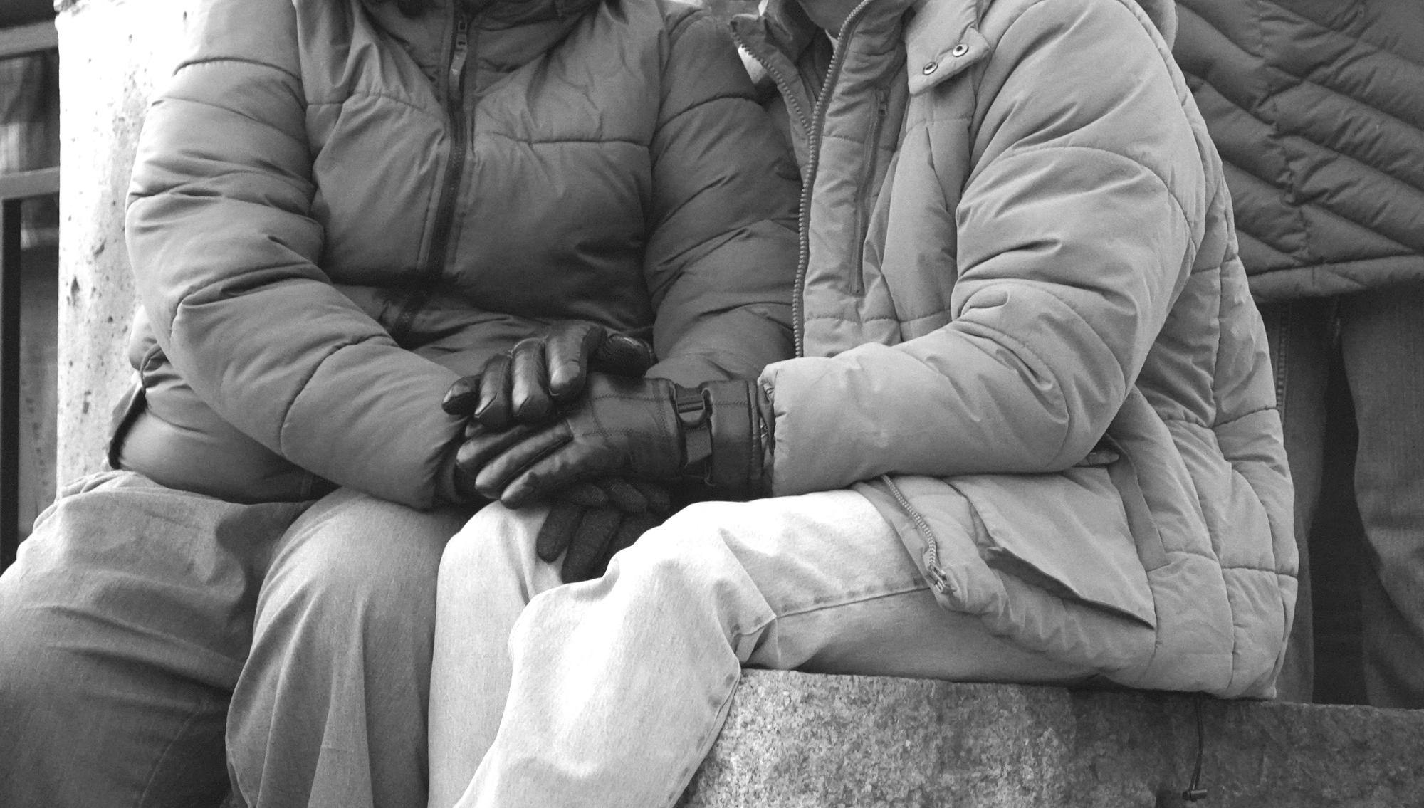 Black-and-white photo of two people, dressed in coats and gloves, seated close to each other with their hands layered together.