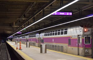 A purple and grey commuter rail train sits in an underground tunnel next to a concrete platform, illuminated by bright fluorescent lights.