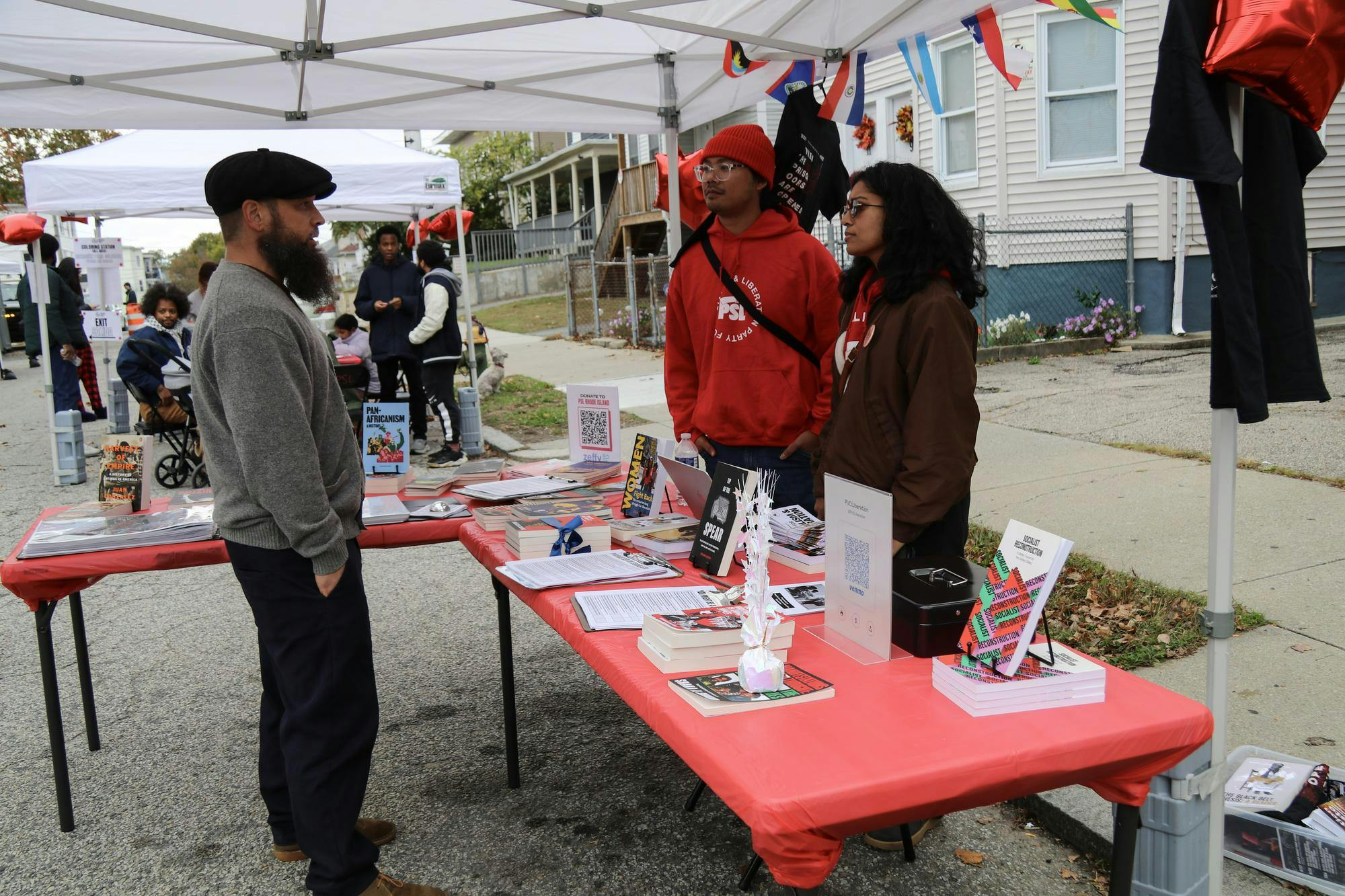 Photo of a person wearing a gray sweater standing in front of a red table with books laid on top talking to a girl wearing a brown sweatshirt and a man wearing a red hoodie on the other side of the table. 