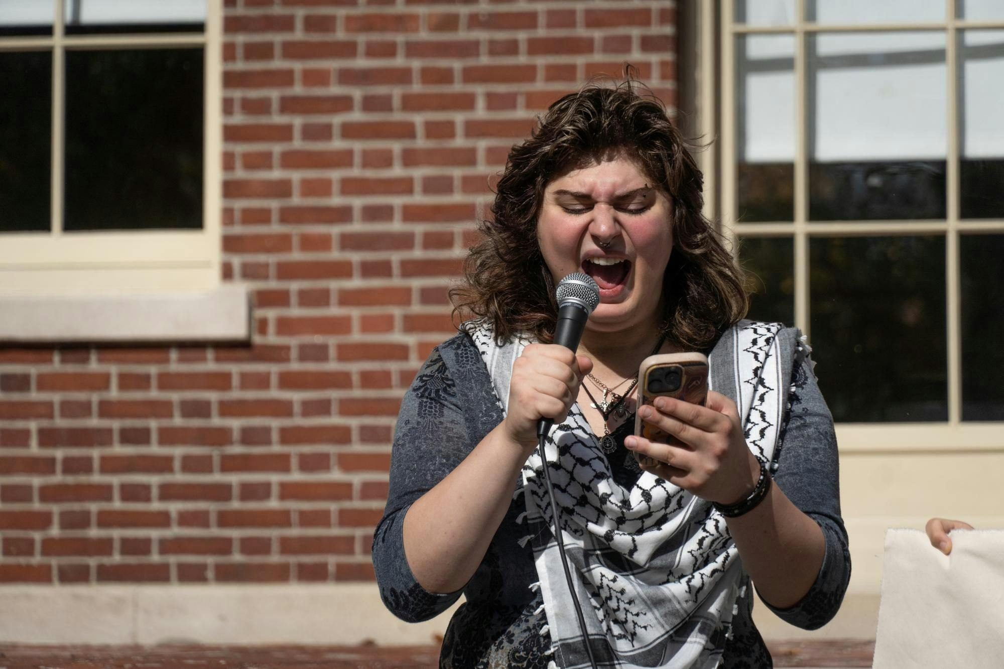 Photo of a woman wearing a keffiyeh, holding a phone and yelling into a microphone.