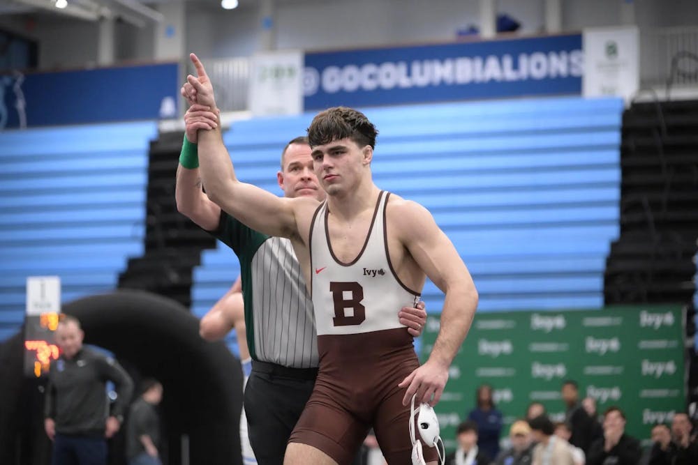 Brown wrestler pointing his finger up with a referee standing behind him and holding him by his waist and wrist.