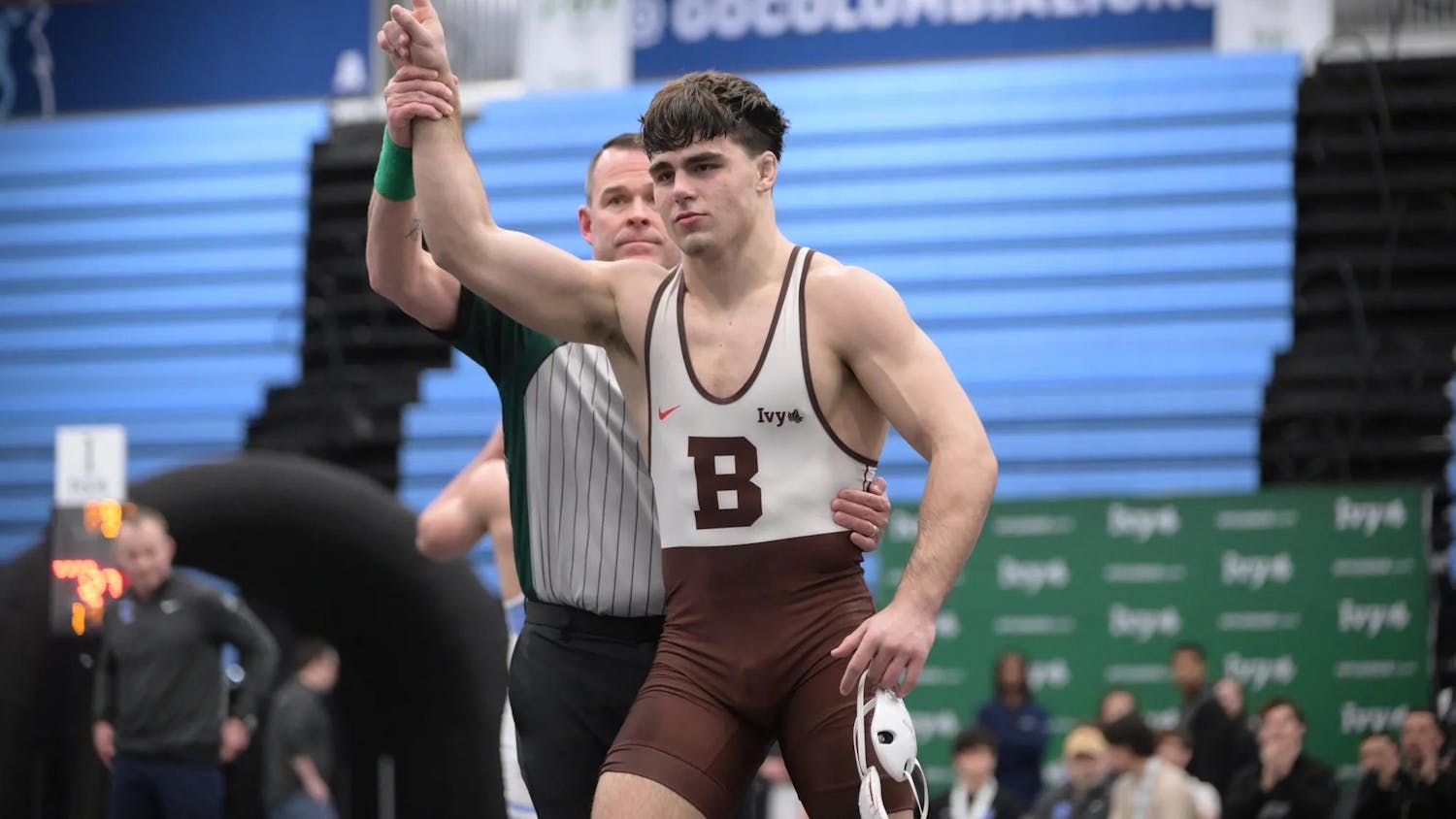 Brown wrestler pointing his finger up with a referee standing behind him and holding him by his waist and wrist.