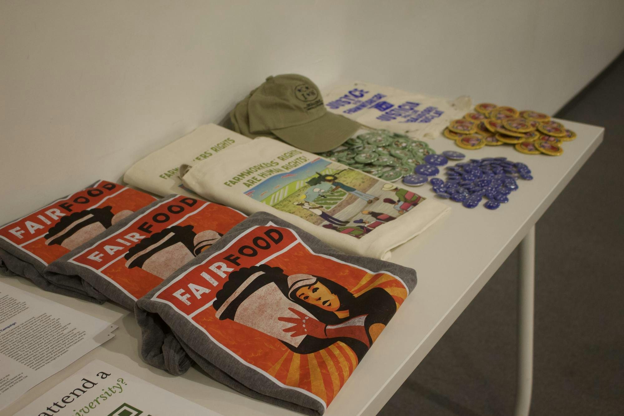 Photo of gray shirts with an illustration of a woman carrying a basket and “FAIRFOOD” label printed on them, white tote bags featuring an illustration and “FARMWORKERS’ RIGHTS ARE HUMAN RIGHTS” label, green caps and various badges, all laid out on a white table.