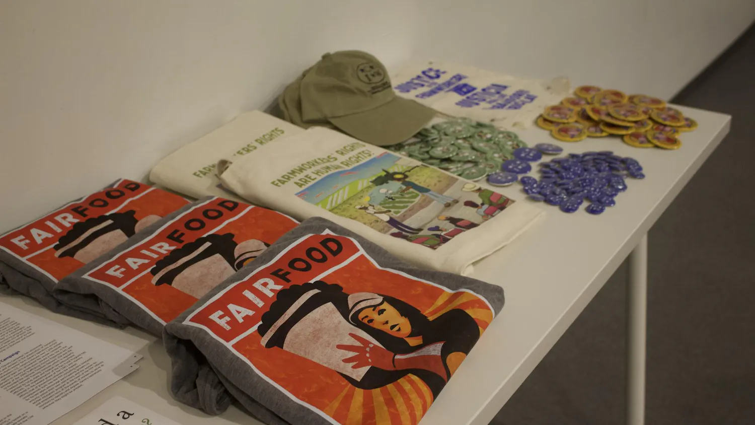 Photo of gray shirts with an illustration of a woman carrying a basket and “FAIRFOOD” label printed on them, white tote bags featuring an illustration and “FARMWORKERS’ RIGHTS ARE HUMAN RIGHTS” label, green caps and various badges, all laid out on a white table.