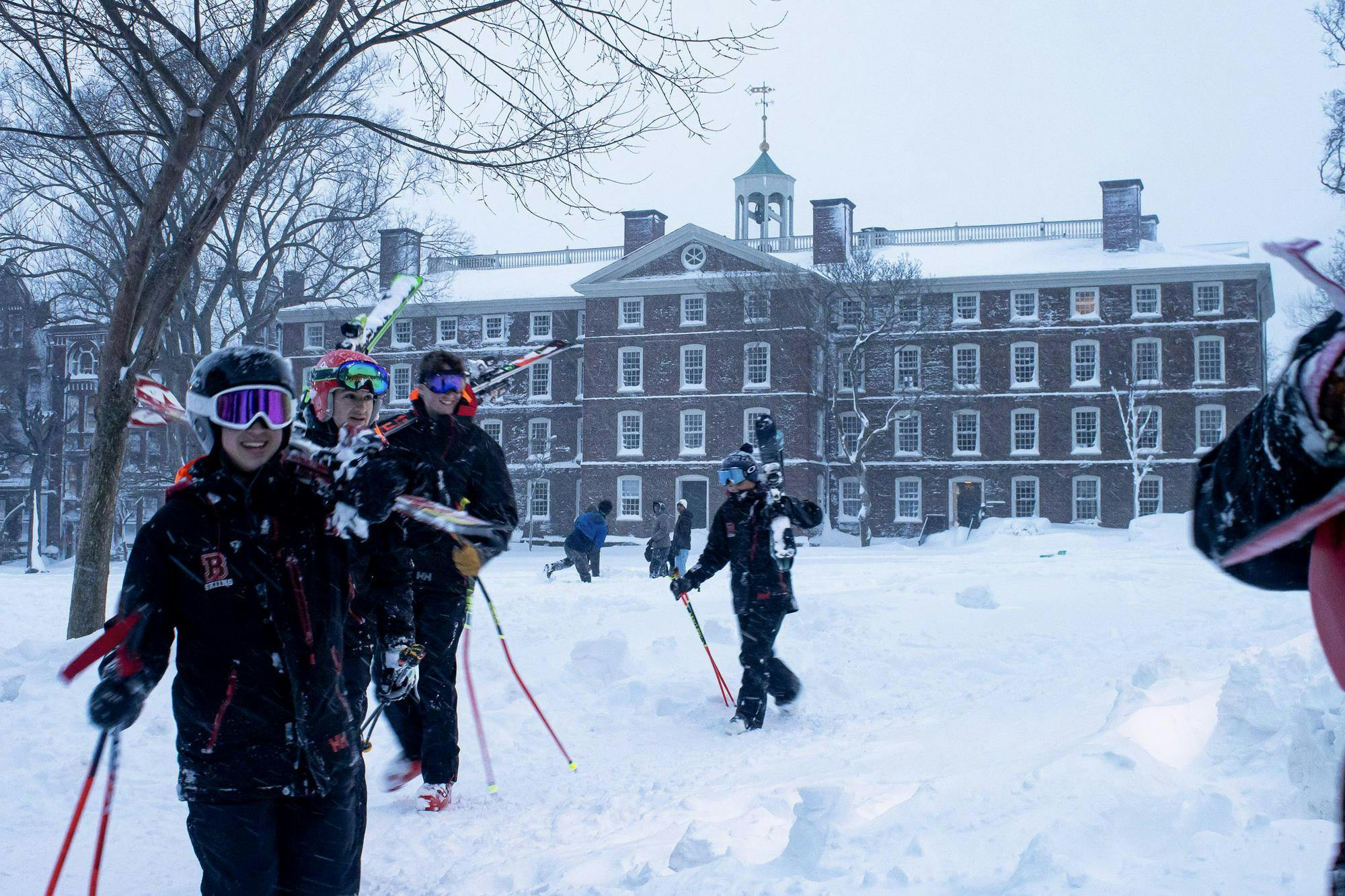 A photo of people wearing ski gear and carrying their skis on the Main Green during a snowstorm. 