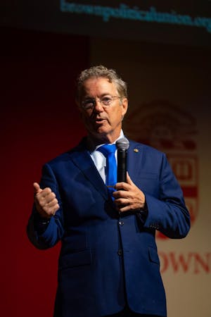 U.S. Sen. Rand Paul, a gray-haired man wearing glasses and a navy blue suit, stands in a spotlight on a stage, speaking into a microphone. 
