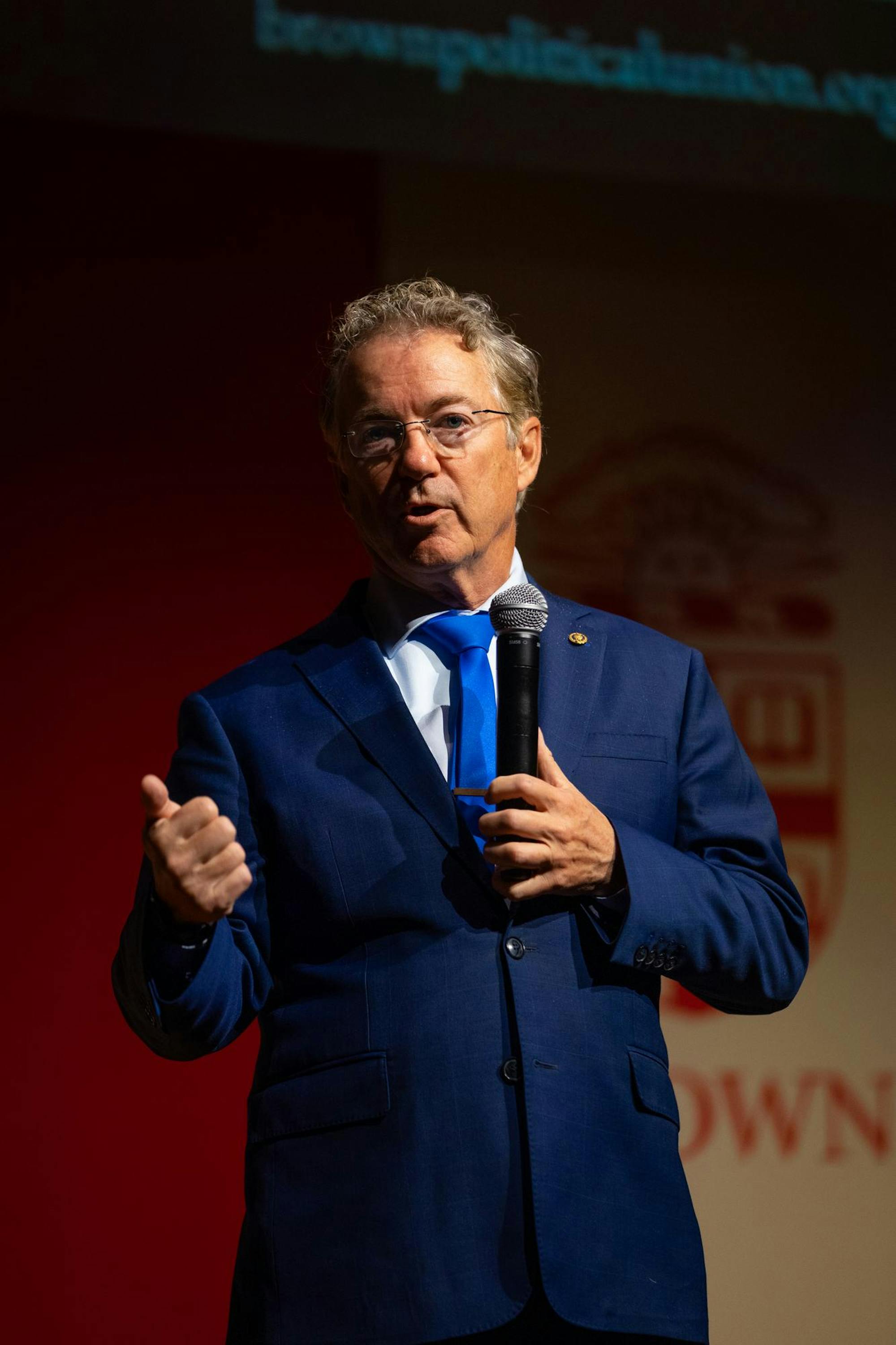 U.S. Sen. Rand Paul, a gray-haired man wearing glasses and a navy blue suit, stands in a spotlight on a stage, speaking into a microphone. 