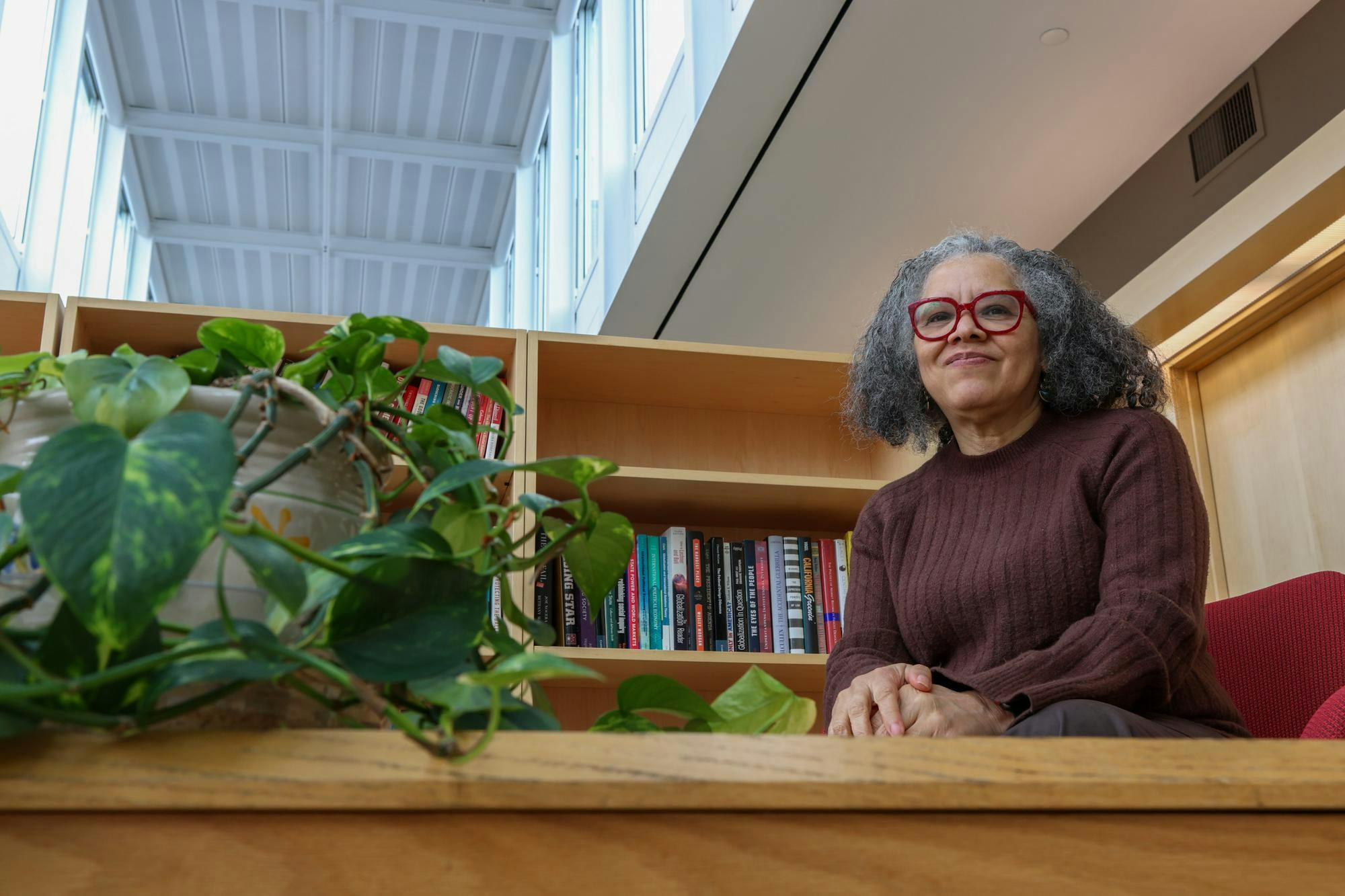 Professor of Political Science Katherine Tate smiles during an interview with The Herald. She wears a brown sweater and bright red glasses frames, and she sits near a bookshelf and potted plant. 