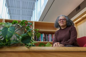 Professor of Political Science Katherine Tate smiles during an interview with The Herald. She wears a brown sweater and bright red glasses frames, and she sits near a bookshelf and potted plant. 