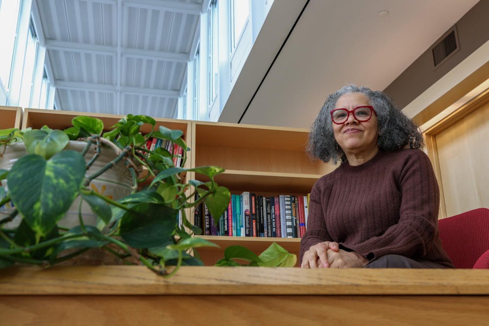 Professor of Political Science Katherine Tate smiles during an interview with The Herald. She wears a brown sweater and bright red glasses frames, and she sits near a bookshelf and potted plant. 
