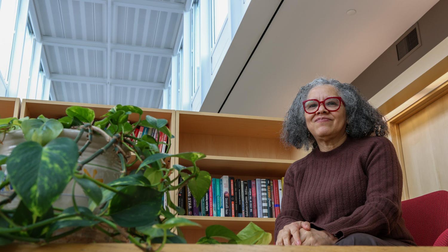 Professor of Political Science Katherine Tate smiles during an interview with The Herald. She wears a brown sweater and bright red glasses frames, and she sits near a bookshelf and potted plant.
