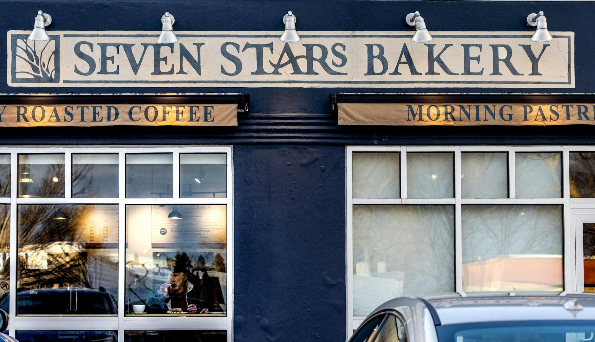 Photo of a blue storefront with a long banner at the top for Seven Stars Bakery and two banners advertising roasted coffee and morning pastries. Two windows allow for a view into the bakery.