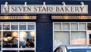 Photo of a blue storefront with a long banner at the top for Seven Stars Bakery and two banners advertising roasted coffee and morning pastries. Two windows allow for a view into the bakery.