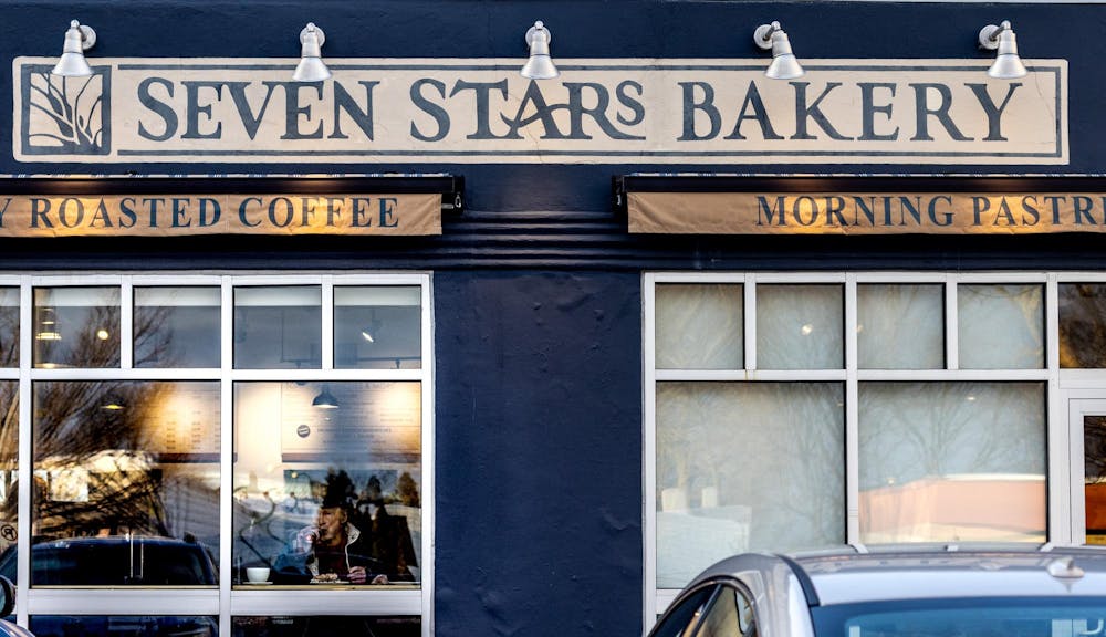 Photo of a blue storefront with a long banner at the top for Seven Stars Bakery and two banners advertising roasted coffee and morning pastries. Two windows allow for a view into the bakery.