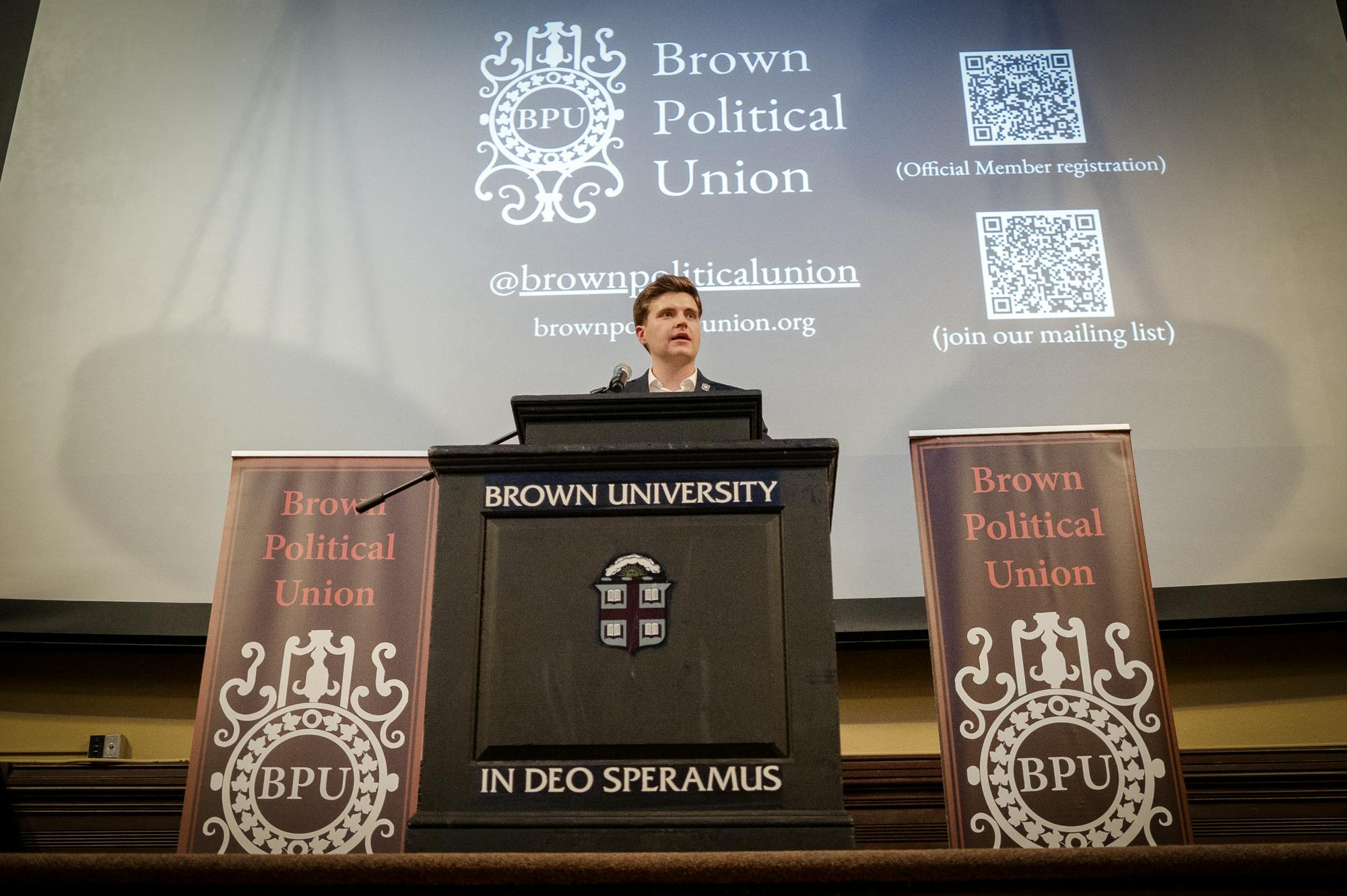 A man in a dark blue suit stands behind a black podium flanked by two brown banners that say "Brown Political Union" in white text.