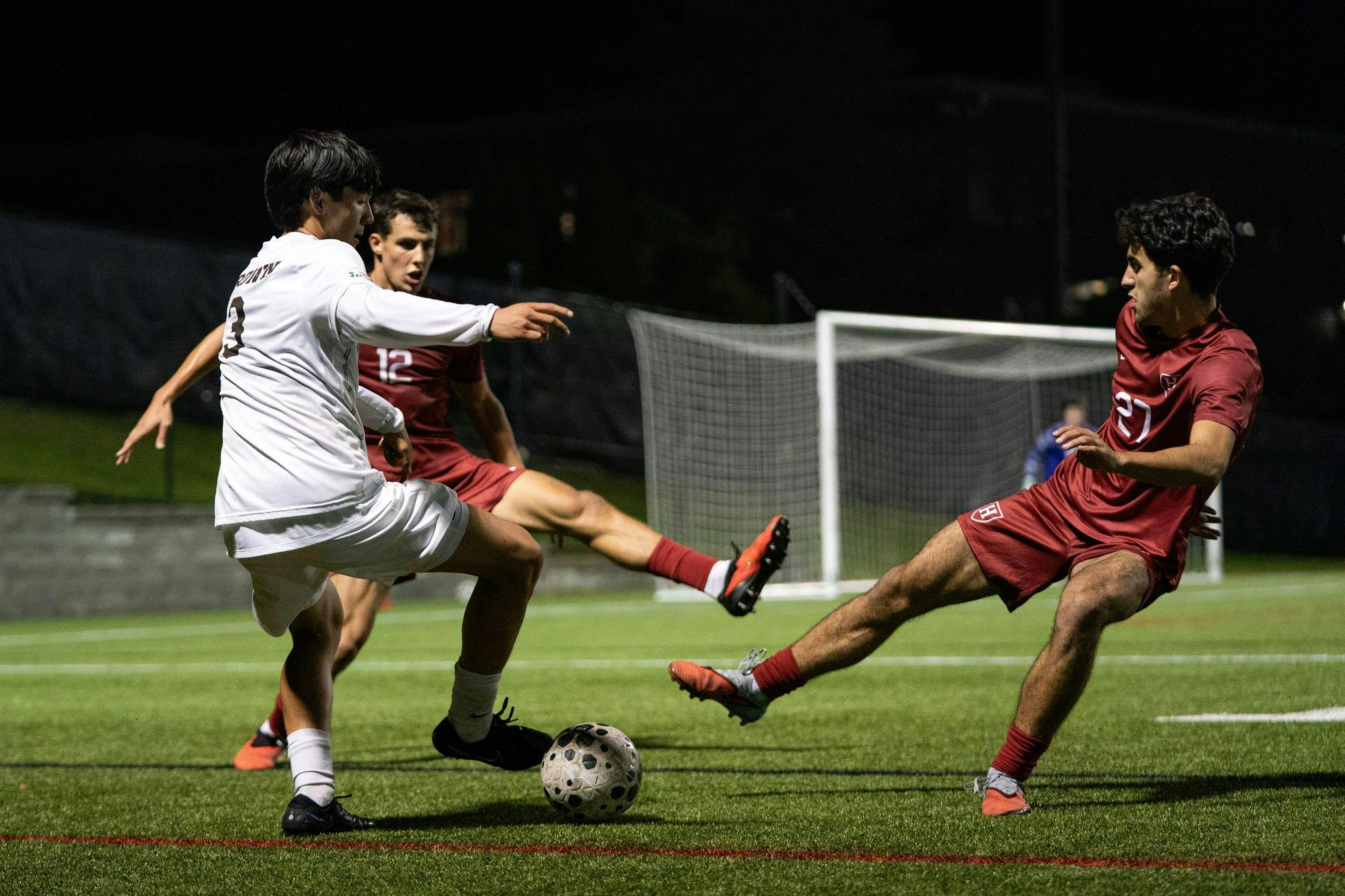 Photo of a Brown soccer player wearing a white uniform dribbling a soccer ball on the field at night as two Harvard players wearing red uniforms kick their legs towards the ball. 