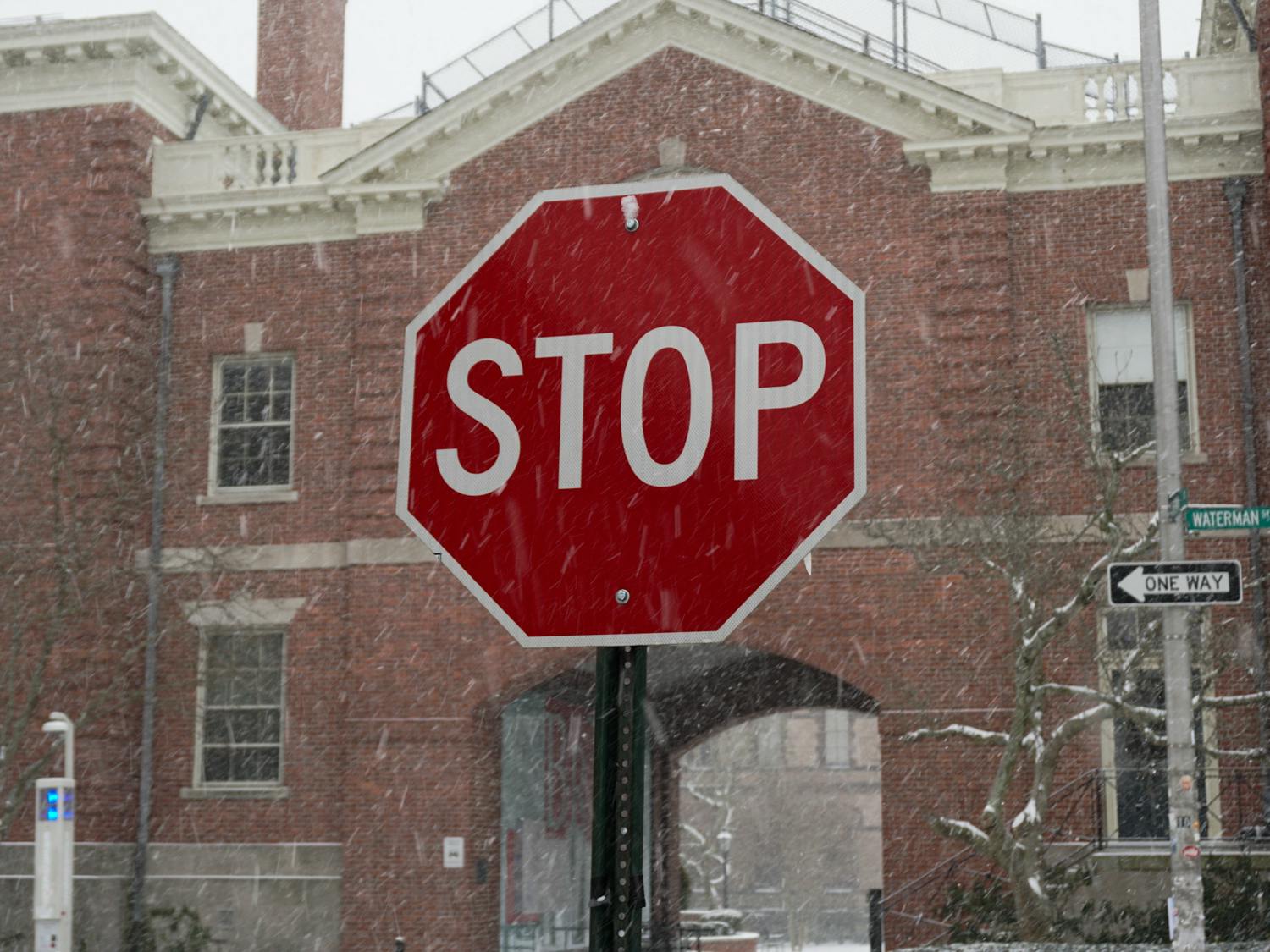 Photo of a stop sign with snow falling around it. The sign is in front of a red brick building.