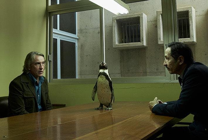A penguin stands on a wooden table between two men, cast by yellow lighting. 