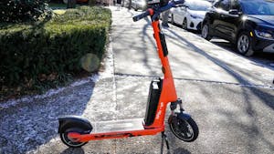 An orange Spin electric scooter is parked on a sidewalk corner lined with specks of snow. A bush and parked cars are in the background.