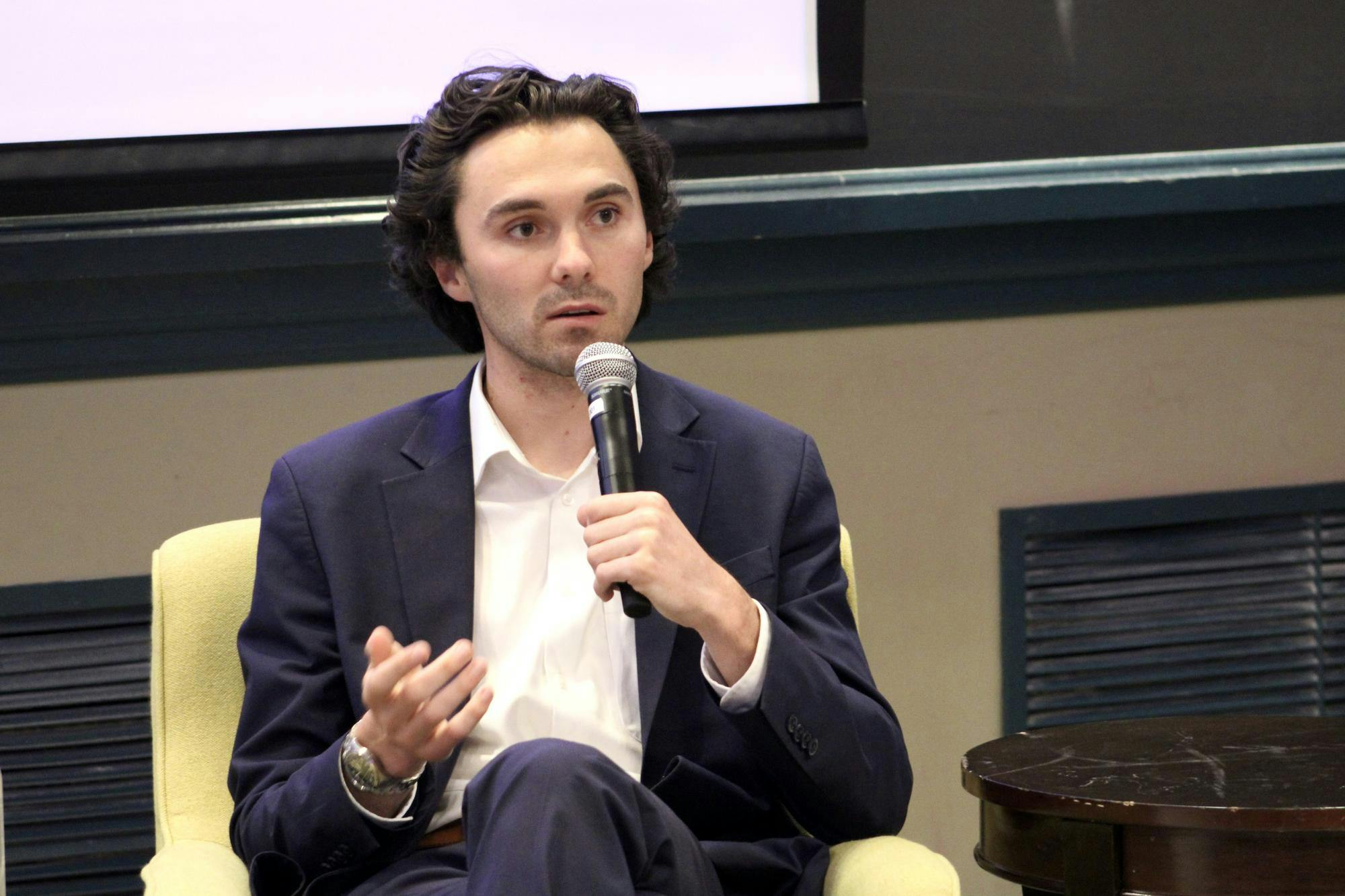 David Hogg sits wearing a navy suit holding a microphone. 