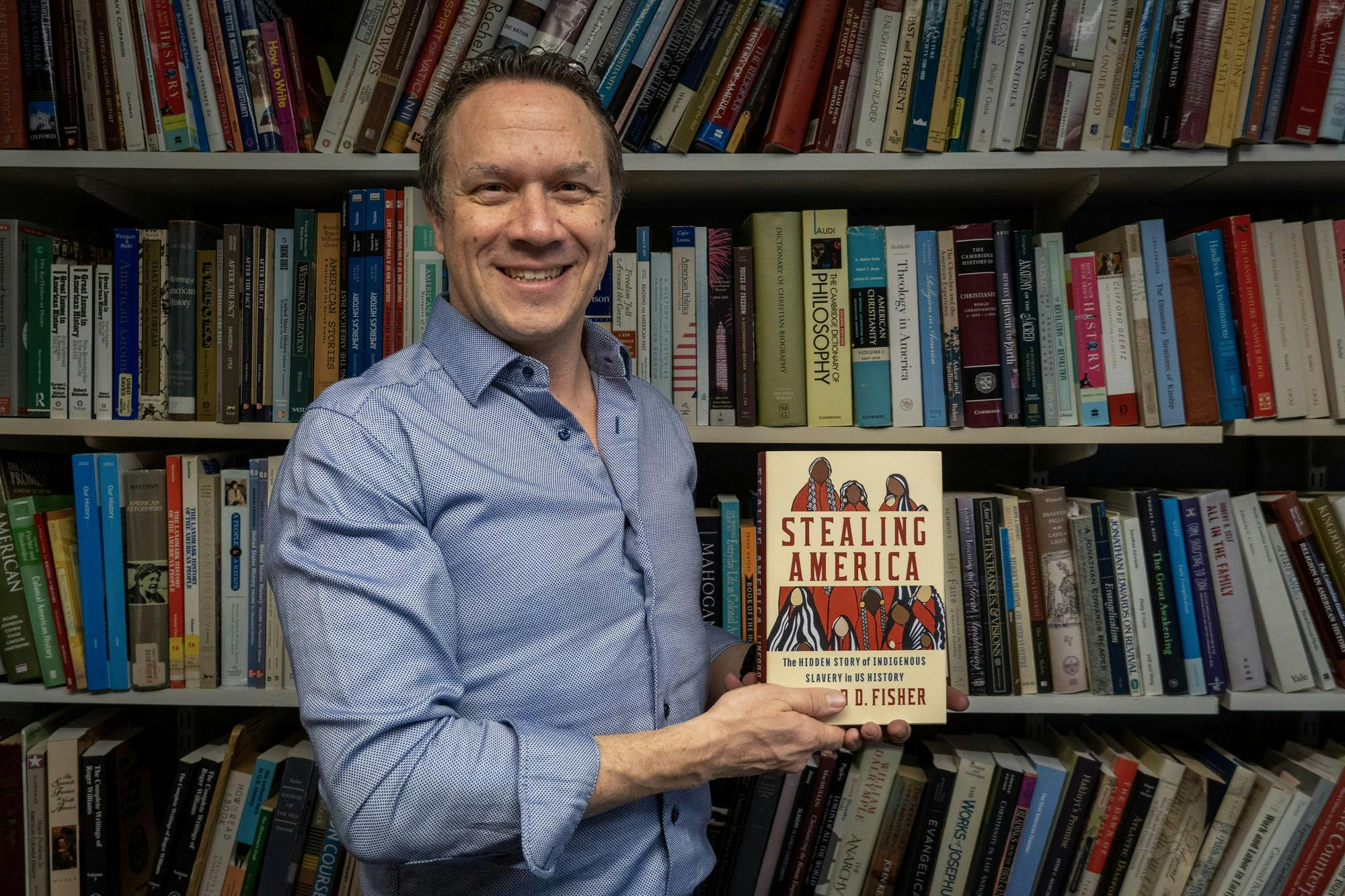 A photo of Professor Linford D. Fisher holding his book “Stealing America” in front of a bookshelf. 