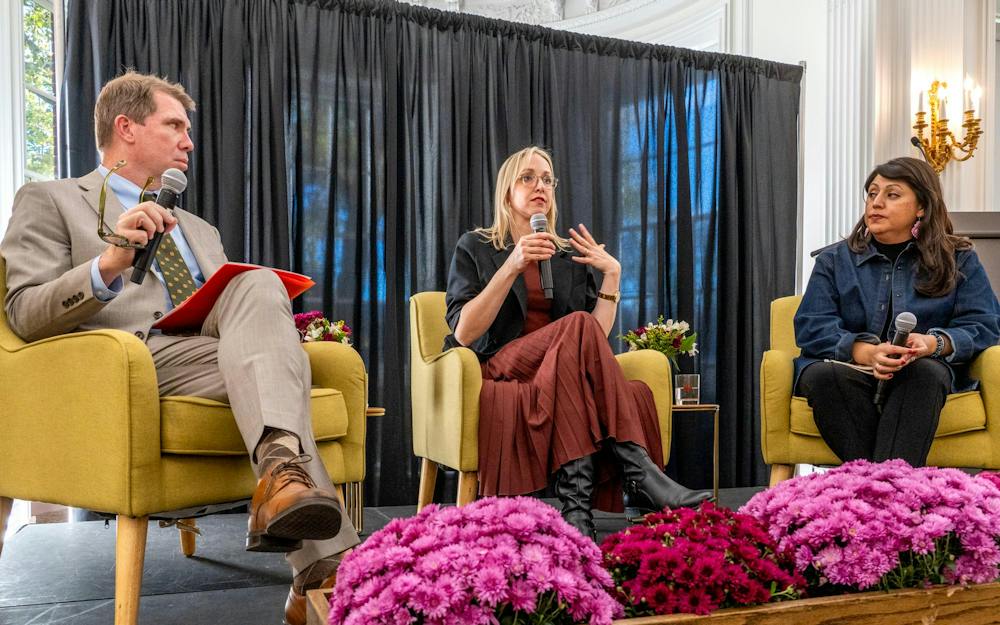 A photo of a man wearing a beige suit next to a woman wearing a black jacket and terracotta dress next to a woman in black pants and a denim jacket all sitting in identical yellow chairs holding microphones.