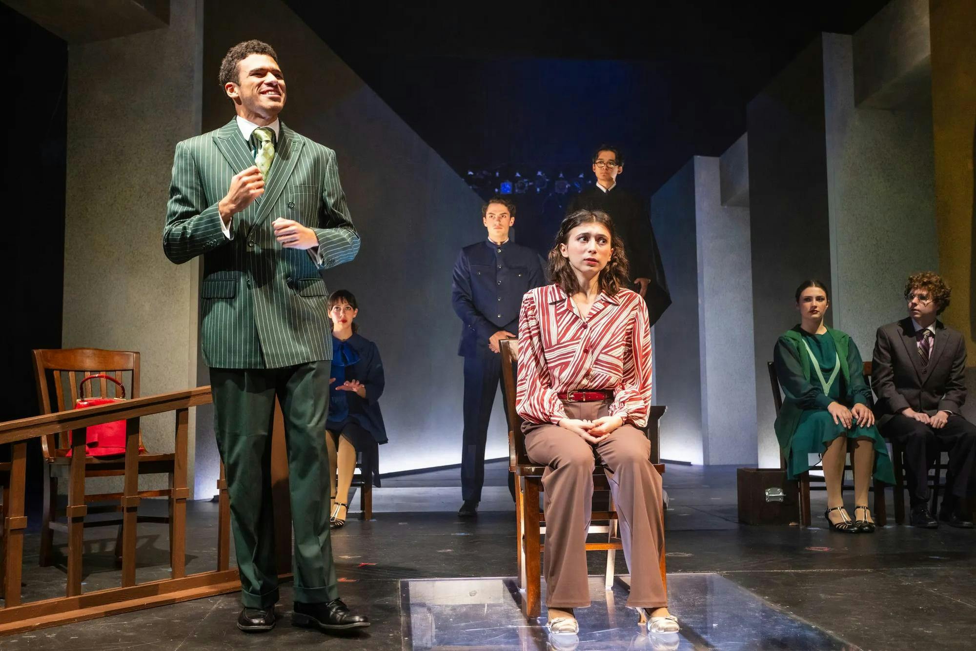A young woman (actor) sits on a stool surrounded by other actors
