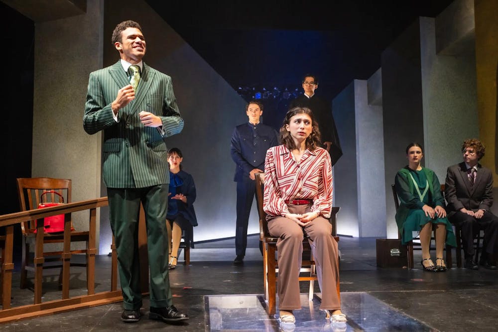 A young woman (actor) sits on a stool surrounded by other actors