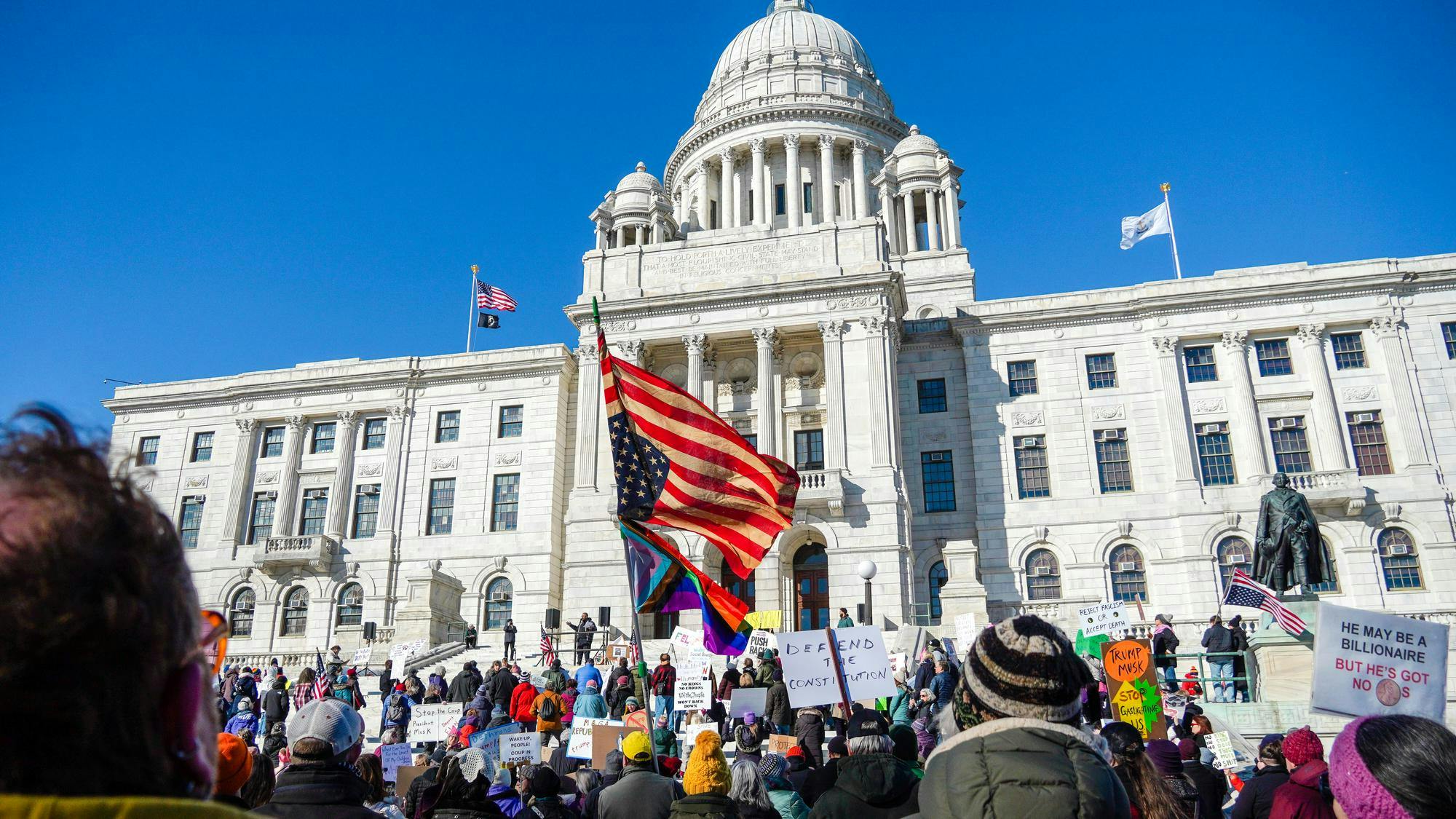 Picture of the rally opposing Elon Musk and the Trump administration outside of the Rhode Island State House. Protestors are waving flags and holding signs.